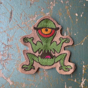 Op de afbeelding: Geborduurde patch met een groen, eenogig wezen met een groot oranje oog, rode omtrek en een roze mond. De patch heeft een bruine rand en een cartoonontwerp. Geschikt voor kleding of accessoires.