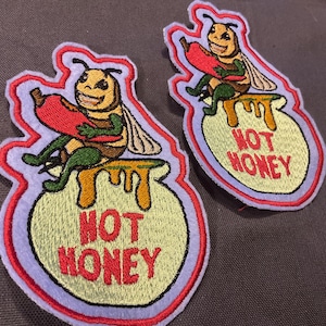 May include: Two embroidered patches with a cartoon bee holding a red chili pepper, sitting on a honey pot. The patches have a light purple border and the words "HOT HONEY" in red.