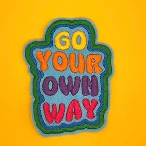 May include: Embroidered patch with the phrase "GO YOUR OWN WAY" in colourful, stitched letters. The patch has a light blue background with a green and blue border, set against a yellow background.