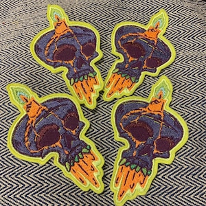 Puede incluir: Cuatro parches bordados con un diseño de calavera con una vela encima. Las calaveras son moradas con detalles naranjas y verdes, delineadas en verde brillante. Los parches están sobre un fondo azul y blanco estampado.