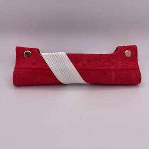 Puede incluir: Un estuche de tela roja con una correa diagonal blanca. El estuche rectangular tiene extremos redondeados, asegurados por remaches dorados. La correa blanca envuelve el estuche, agregando un elemento de contraste.