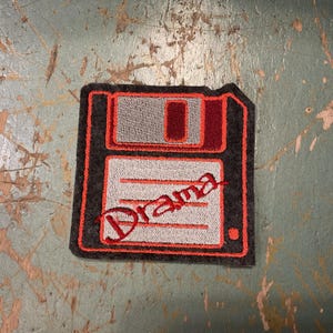 May include: Embroidered patch featuring a retro floppy disk design. The patch is gray with a red and orange outline, and the word "Drama" is stitched across the front in red thread. A fun, nostalgic accessory.