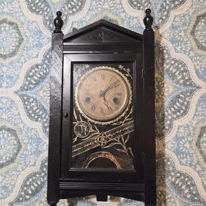 May include: A black wooden wall clock with a pointed top and finials. The clock face features Roman numerals and ornate gold-toned floral detailing on the glass door. A central monogram with the letters GC is visible below the clock face.