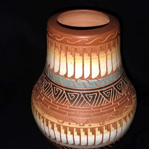 May include: A terracotta clay vase featuring a wide, rounded body and a narrow neck. The surface is decorated with etched geometric patterns, including vertical lines, a teal wave band, and black triangles, accented with white and tan gradients.