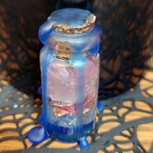 Op de afbeelding: Een kleine glazen fles met een kurk, verzegeld met blauwe was. De fles bevat roze en rode gedroogde bloemen. De was is langs de zijkanten van de fles naar beneden gedruppeld, wat een decoratief effect creëert.