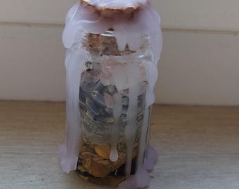 May include: A small glass bottle with a cork stopper, sealed with light purple wax. The bottle contains various dried herbs and crystals. The wax has dripped down the sides of the bottle. The bottle is sitting on a wooden surface.