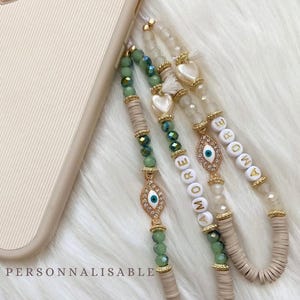 Könnte beinhalten: Perlen-Handyketten in Grün, Weiß und Beige. Sie haben goldfarbene Akzente, Anhänger mit bösen Augen und das Wort "AMORE" in weißen Perlen. Der Text "PERSONNALISABLE" ist auf der Handyhülle zu sehen.