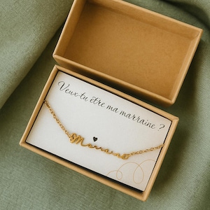 Peut inclure: Un collier doré avec le mot "Marraine" en écriture cursive, présenté dans une petite boîte en carton ouverte. Le collier est posé sur une carte blanche avec la phrase française "Veux-tu être ma marraine ?" imprimée en noir.
