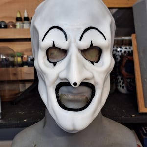 May include: White plastic mask with black painted details, including eyebrows, eyes, and a mouth. The mask has a realistic, slightly grotesque appearance.