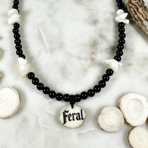 May include: A black beaded necklace with white accents and a pendant that reads "Feral". The necklace is displayed on a white marble surface, surrounded by pieces of antler.