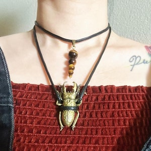 May include: A layered necklace with a gold beetle pendant and a brown and gold tiger's eye stone pendant.