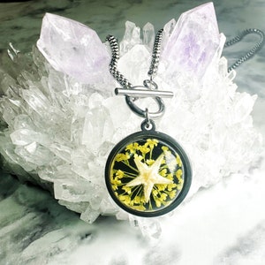 May include: A silver chain necklace with a toggle clasp and a round pendant. The pendant is filled with clear resin and contains a dried starfish and yellow flowers.