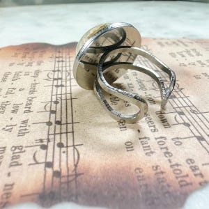 May include: A silver-toned adjustable ring featuring a circular setting with a printed musical score background. The band is open and split into two thin, curved metal wires that wrap around the finger, showcasing a metallic finish.