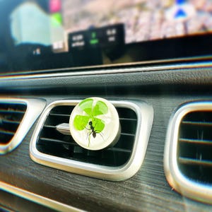 May include: A clear plastic car air freshener with a four-leaf clover and a black ant inside. The air freshener is attached to a car's air vent.