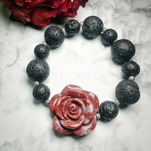 May include: A beaded bracelet featuring large, textured black lava rock beads and a central red rose-shaped stone. The bracelet is strung with silver-colored spacers. The rose has a marbled appearance.
