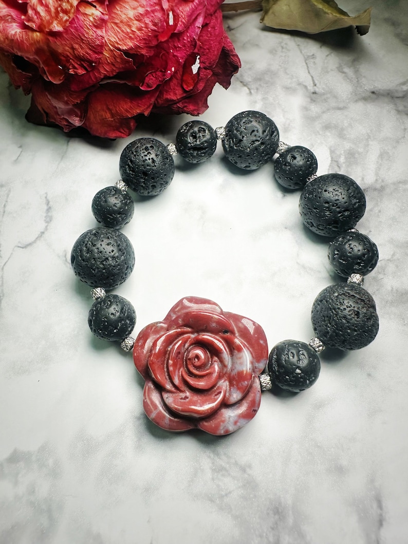 May include: A beaded bracelet featuring large, textured black lava rock beads and a central red rose-shaped stone. The bracelet is strung with silver-colored spacers. The rose has a marbled appearance.