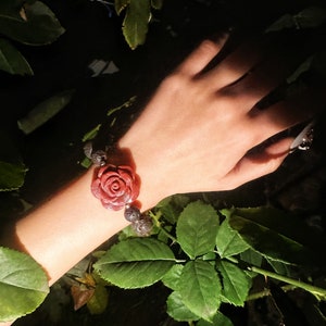 May include: A red rose-shaped bracelet with dark gray beads. The bracelet is worn on a wrist, surrounded by green leaves and a dark background. The rose is the focal point, with detailed petals.