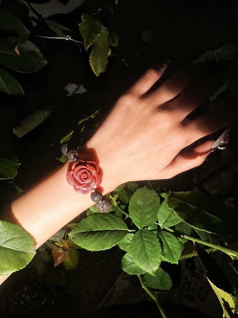 May include: A red rose-shaped bracelet with dark gray beads. The bracelet is worn on a wrist, surrounded by green leaves and a dark background. The rose is the focal point, with detailed petals.