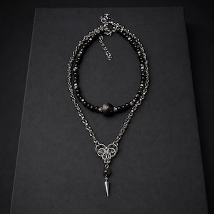 May include: A layered necklace featuring a top strand of black beads with a central dark stone and a bottom silver-toned chain strand. The lower chain includes a complex woven metal link design, a single dark bead, and a pointed silver-toned spike pendant.