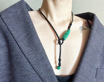May include: A black cord necklace with a green stone pendant. The pendant is oval shaped and has a marbled pattern. The necklace is worn by a person with a gray blazer.