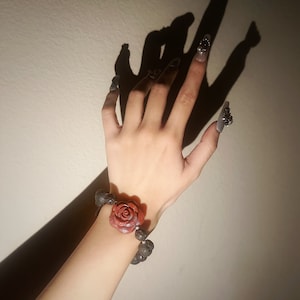 May include: A bracelet featuring a large, red rose centerpiece and dark gray beads. The bracelet is worn on a wrist with long, gray nails and decorative nail art. The image is lit with a strong light source, casting a shadow.