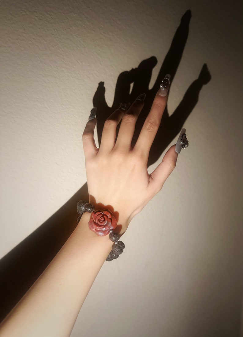 May include: A bracelet featuring a large, red rose centerpiece and dark gray beads. The bracelet is worn on a wrist with long, gray nails and decorative nail art. The image is lit with a strong light source, casting a shadow.