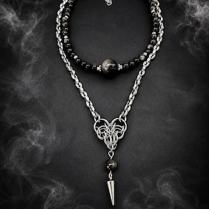 May include: A layered necklace featuring a strand of dark, polished spherical beads and a silver-toned metal chain. The chain incorporates an intricate woven metal link design, a single dark bead, and a pointed, cone-shaped silver metal pendant.