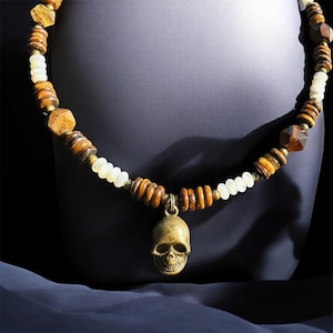 May include: A necklace featuring a skull pendant and a beaded design. The necklace has a mix of brown, white, and gold-colored beads, with a skull charm hanging in the center. The beads are various shapes and sizes.