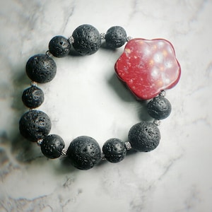 May include: A black lava stone bead bracelet with a red, flower-shaped centerpiece. The bracelet features round, porous black beads strung together with silver-colored wire. The centerpiece is a deep red color with lighter speckles.