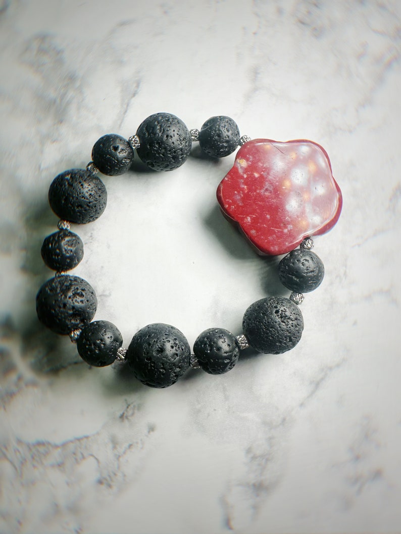 May include: A black lava stone bead bracelet with a red, flower-shaped centerpiece. The bracelet features round, porous black beads strung together with silver-colored wire. The centerpiece is a deep red color with lighter speckles.