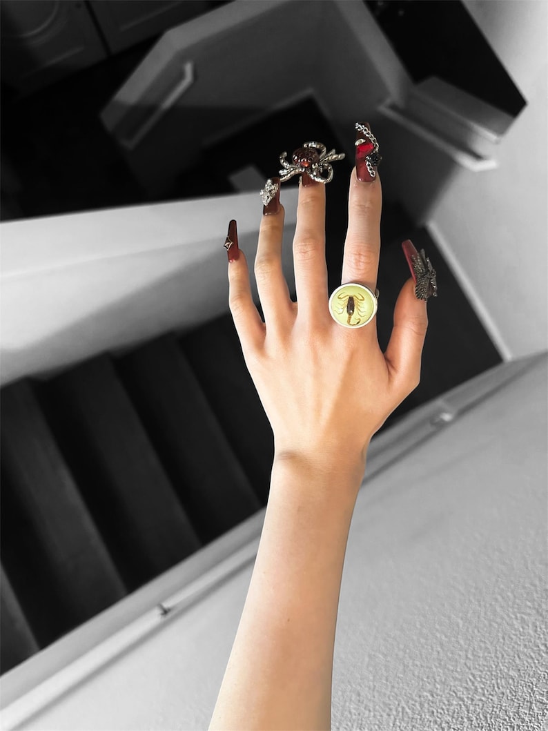 May include: A round ring featuring a yellow scorpion graphic on the center finger, accompanied by a silver-toned spider ring on the index finger and a dark butterfly-shaped ring on the pinky finger, worn with long dark red manicured nails.