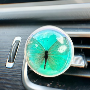 May include: A round, clear car air freshener with a turquoise butterfly design. The air freshener is attached to a car's air vent.