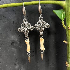 May include: A pair of silver-toned earrings with a chainmail design, bone-shaped charms, and gold-colored cone pendants. The earrings are suspended from a green stem against a dark background.