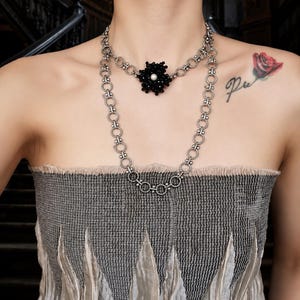 May include: A double-layered silver-toned chain necklace featuring a central floral-shaped pendant made of small black beads with a single white pearl at the center. The necklace consists of interconnected circular links with a textured finish.