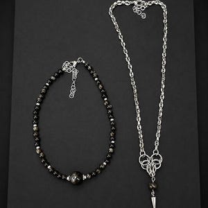 May include: A beaded necklace featuring dark, reflective spherical beads with silver-toned spacers and a larger central bead, alongside a silver-toned chain necklace with a complex woven metal pendant and a single dangling spike-shaped charm.