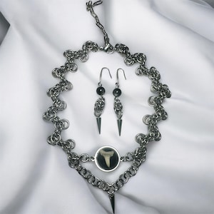 May include: A silver-toned necklace and earring set. The necklace features a chainmail design with a pendant containing a shark tooth. The earrings have black beads and cone-shaped dangles. The jewelry is displayed on a white background.