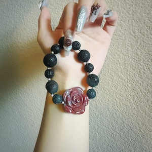 May include: A black lava bead bracelet with a red rose-shaped centerpiece and dark green beads. The bracelet is held up against a neutral background.