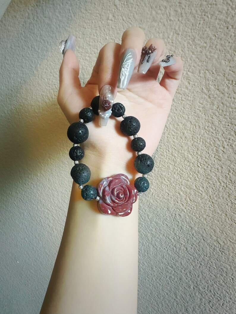 May include: A black lava bead bracelet with a red rose-shaped centerpiece and dark green beads. The bracelet is held up against a neutral background.