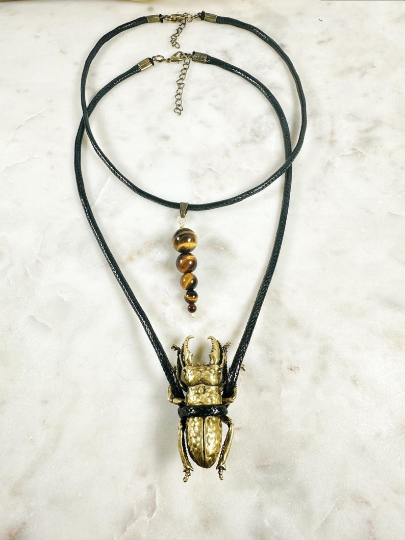 May include: A gold-toned beetle pendant hangs from a black leather cord necklace. The pendant is detailed with a textured surface and is tied with a black cord. A second black leather cord necklace with a tiger's eye bead pendant hangs above the beetle pendant.