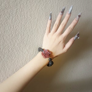 May include: A rose-themed bracelet with a dark red rose and black beads. The bracelet is worn on a wrist with long, gray, decorated fingernails. The background is a textured, light beige wall.