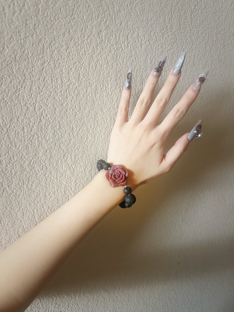 May include: A rose-themed bracelet with a dark red rose and black beads. The bracelet is worn on a wrist with long, gray, decorated fingernails. The background is a textured, light beige wall.