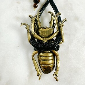May include: A gold-colored beetle pendant with black accents hangs from a black leather cord. The pendant is detailed with intricate carvings and a textured surface.