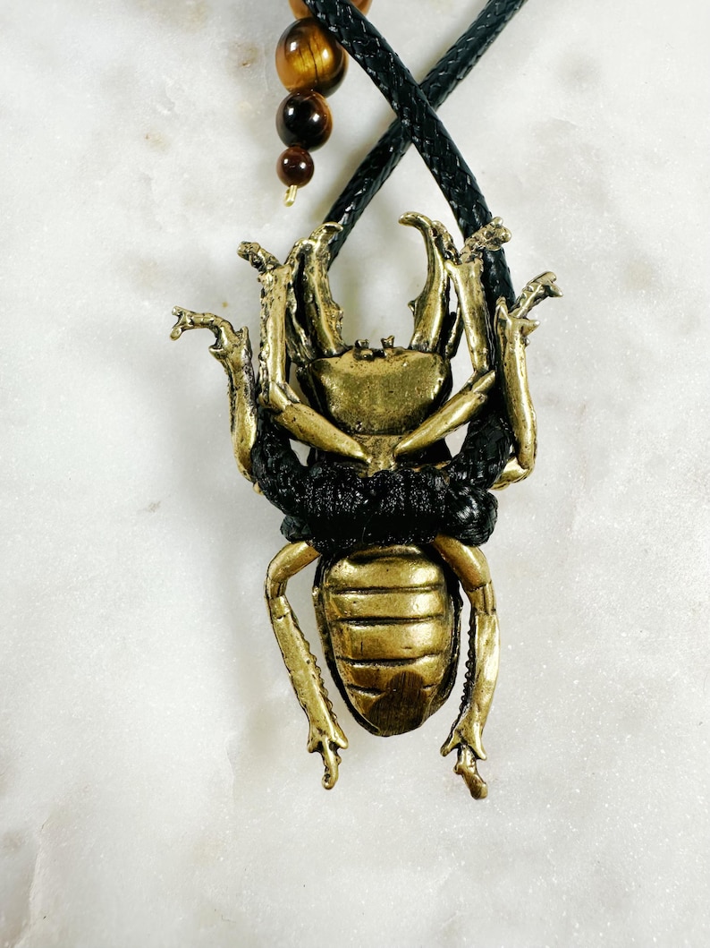 May include: A gold-colored beetle pendant with black accents hangs from a black leather cord. The pendant is detailed with intricate carvings and a textured surface.