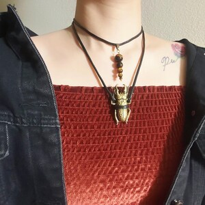 May include: A person wearing a red velvet top with a black choker necklace and a long necklace with a gold beetle pendant and brown tiger eye beads.