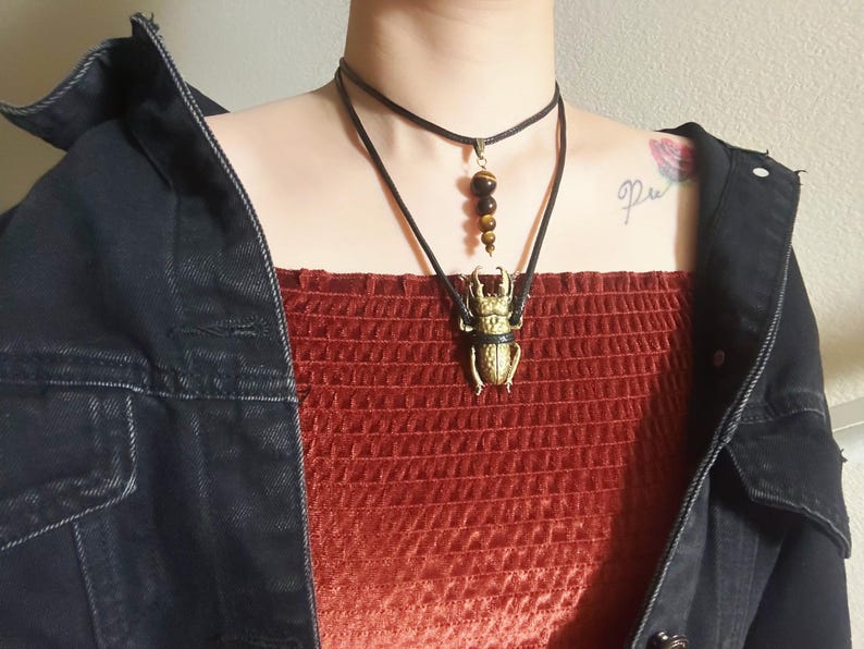 May include: A person wearing a red velvet top with a black choker necklace and a long necklace with a gold beetle pendant and brown tiger eye beads.