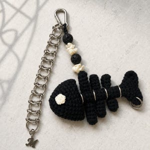 May include: A keychain featuring a black crocheted fish skeleton with a white rose accent on the head. The design includes a silver-toned metal chain, a lobster clasp, a letter M charm, and a strand of black lava beads and white shell-like beads.