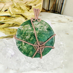 May include: A green and white agate stone pendant with a copper wire wrap. The pendant is sitting on a bed of clear quartz crystals.