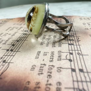 May include: A silver-toned metal ring featuring a dome-shaped, pale yellow resin cabochon. Encased within the resin is a dark brown scorpion specimen. The ring band is constructed from two thin, curved metal wires that meet at the setting.