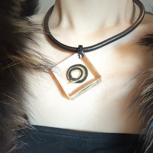 May include: A square pendant necklace with a clear resin cube containing a small black and white snake. The necklace is made of brown leather cord.