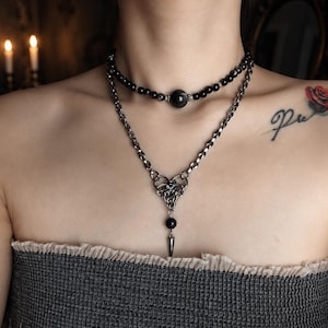 May include: A layered necklace set featuring a choker made of small black beads with a central larger bead and a longer silver-toned chain necklace with a complex woven metal link centerpiece, a single black bead, and a pointed metal pendant.